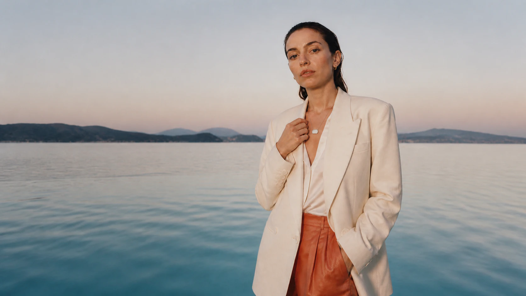 Woman wearing an ivory Presence Key beside open water and distant mountains at dusk